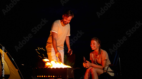 A cute woman and a handsome man are sitting on folding chairs near the tent by the fire, drinking beer and having fun at night on the beach by the sea.