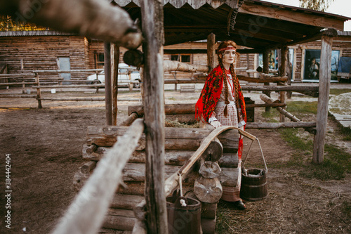 A beautiful Slavic girl with long blonde hair and brown eyes in a white and red embroidered suit is sitting next to a wooden house.Traditional clothing of the Ukrainian region.