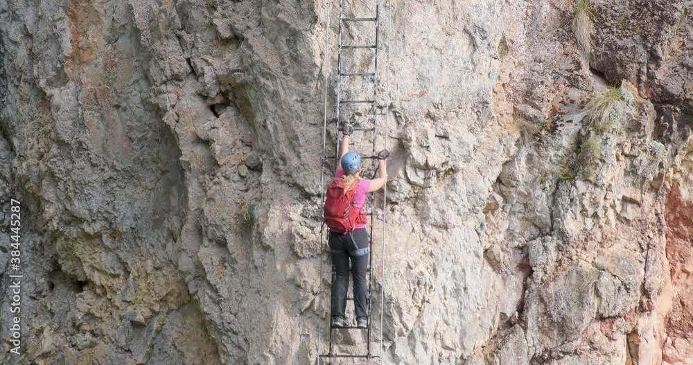 Woman with red backpack climbs a vertical ladder on a via ferrata route called Piatra Soimilor, in Harghita county, Romania. Adventure, climbing, tilt up shot. Woman with red backpack climbs a vertical ladder on a via ferrata route called Piatra Soimilor, in Harghita county, Romania. Adventure, climbing, tilt up shot.