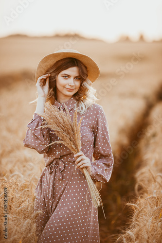 A young girl with long curly hair and a hat , in a brown dress with white polka dots, stands in a wheat field