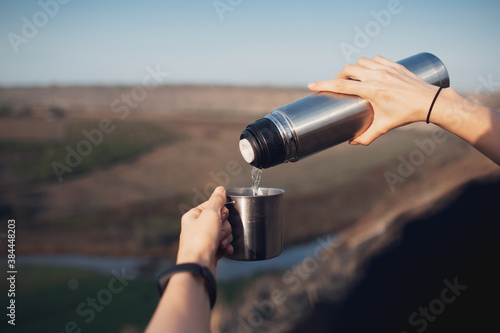 Close-up of female hands, pouring tea into the steel cup from thermos, on outdoor background. Travel concept.