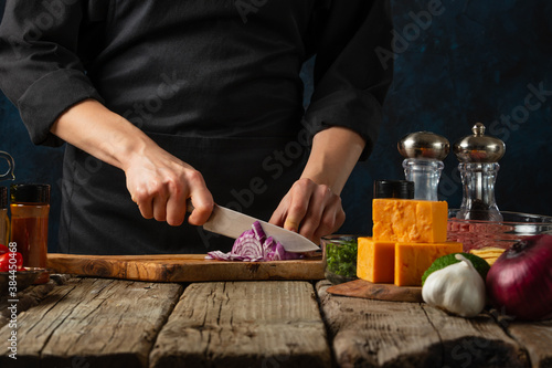 The chef in black uniform cuts with knife onion on wooden chopped board on dark blue background with other ingredients. Backstage of cooking meal on wooden rustic table. Food concept.