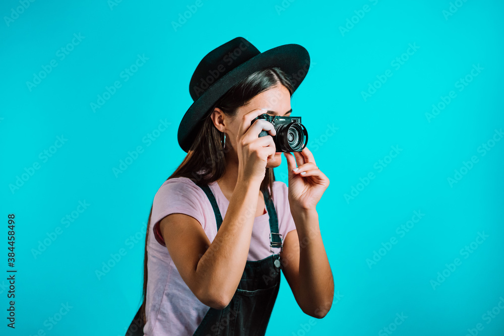 Young pretty woman in overall takes pictures with DSLR camera over blue background in studio. Girl smiling and having fun as photographer. Young pretty woman in overall takes pictures with DSLR camera over blue background in studio. Girl smiling and having fun as photographer.