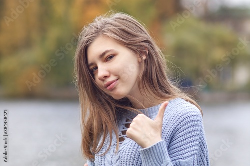 Portrait of young happy positive woman, teenager girl is showing thumb up, like, approval gesture, looking at camera and smiling in autumn park