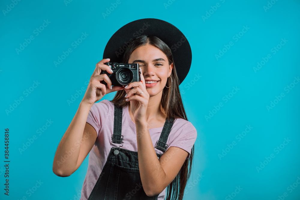 Young pretty woman in overall takes pictures with DSLR camera over blue background in studio. Girl smiling and having fun as photographer. Young pretty woman in overall takes pictures with DSLR camera over blue background in studio. Girl smiling and having fun as photographer.