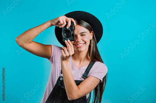 Young pretty woman in overall takes pictures with DSLR camera over blue background in studio. Girl smiling and having fun as photographer. © kohanova1991