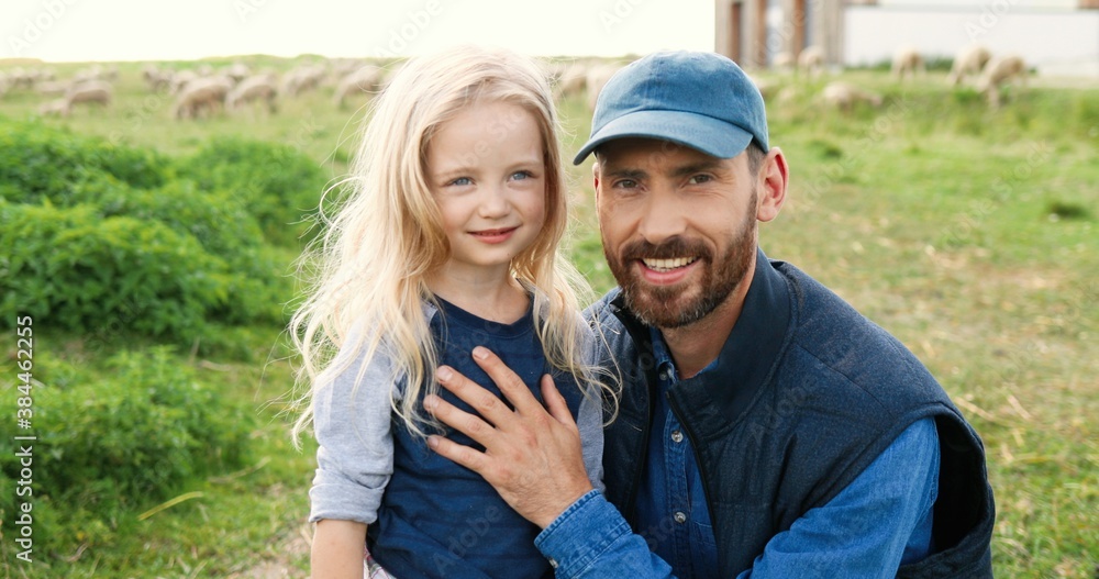 Portrait of Caucasian young handsome father standing outdoor with cute little daughter and smiling to camera in green field. Sheep flock grazing on background a pasture. Smiled man and small girl. Portrait of Caucasian young handsome father standing outdoor with cute little daughter and smiling to camera in green field. Sheep flock grazing on background a pasture. Smiled man and small girl.