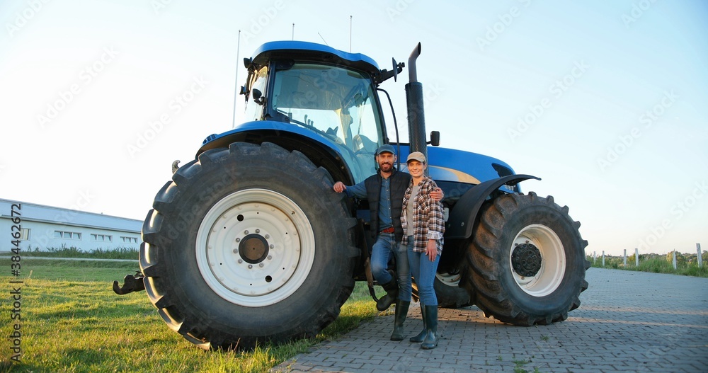 Portrait of Caucasian happy couple of married farmers standing in hugs at tractor, looking at each other and smiling to camera. Outdoor. Man hugging woman. Agricultural vehicle. Wife and husband. Portrait of Caucasian happy couple of married farmers standing in hugs at tractor, looking at each other and smiling to camera. Outdoor. Man hugging woman. Agricultural vehicle. Wife and husband.