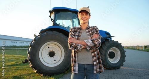 Portrait shot of beautiful Caucasian young woman in cap standing outdoor at farm with big tractor machine on background and smiling joyfully to camera. Pretty happy female farmer in field. Agriculture