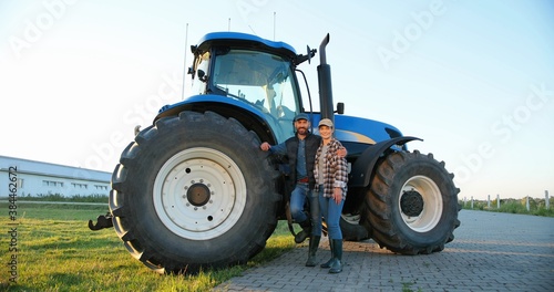 Portrait of Caucasian happy couple of married farmers standing in hugs at tractor, looking at each other and smiling to camera. Outdoor. Man hugging woman. Agricultural vehicle. Wife and husband.