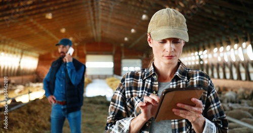 Young Caucasian happy pretty woman in hat using tablet device and smiling in farm stable. Portrait. Female farmer tapping and scrolling on gadget computer in shed. Man speaking on phone on background.