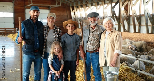 Portrait of happy Caucasian family of three generations standing in shed with livestock and smiling. Old parents with children and grandchildren in stable. farmers at farm. Indoor. Sheep on background