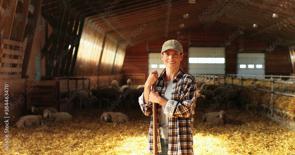 Portrait of beautiful Caucasian young woman in hat looking at camera with smile and leaning on stick of pitchfork. Pretty female shepherd smiling in stable. Dolly shot. Portrait of beautiful Caucasian young woman in hat looking at camera with smile and leaning on stick of pitchfork. Pretty female shepherd smiling in stable. Dolly shot.