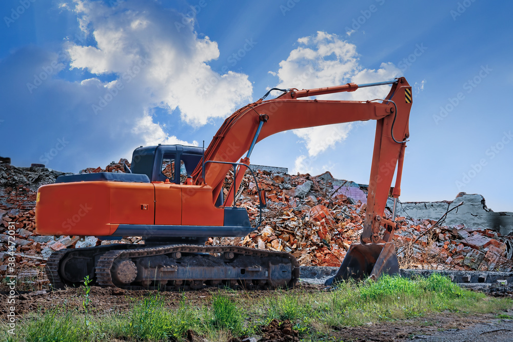 An orange excavator stands against the backdrop of a destroyed building and a blue sky. An orange excavator stands against the backdrop of a destroyed building and a blue sky.