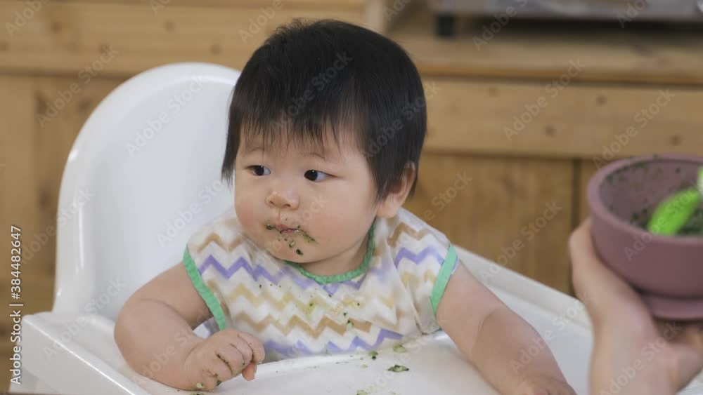 innocent asian young toddler sitting in the baby chair at home is looking away while being fed with pureed food by her mom at breakfast. innocent asian young toddler sitting in the baby chair at home is looking away while being fed with pureed food by her mom at breakfast.
