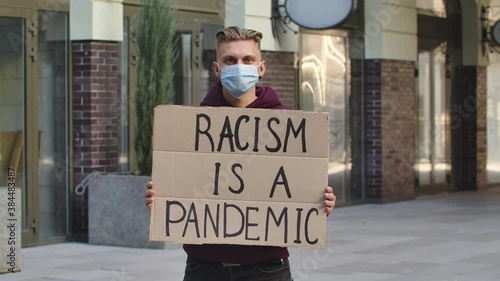 Young man in medical mask stands with a cardboard poster RACISM IS A PANDEMIC in a public place outdoor. No racism, tolerance and political correctness in society. Slow motion. Close up. photo