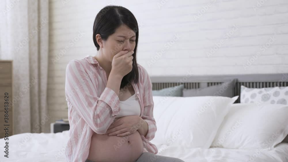 portrait expectant mother sitting in the bedroom is overcame by a wave a nausea and putting her hand on the mouth to avoid vomiting during daytime. portrait expectant mother sitting in the bedroom is overcame by a wave a nausea and putting her hand on the mouth to avoid vomiting during daytime.