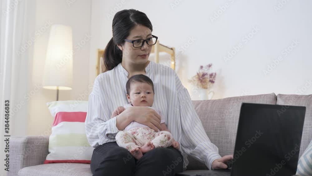 asian businesswoman sitting on the sofa and holding her cute baby is typing on the laptop with concentration at home living room. asian businesswoman sitting on the sofa and holding her cute baby is typing on the laptop with concentration at home living room.