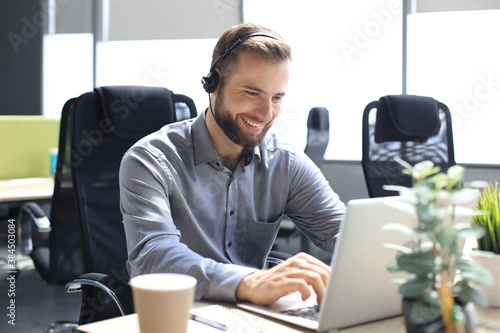Fotografie Smiling male call-center operator with headphones sitting at modern office, consulting online information in a laptop, looking up information in a file in order to be of assistance to the client