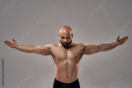 Showing his perfect body. Muscular strong bald man with naked torso standing with outstretched arms isolated over grey background