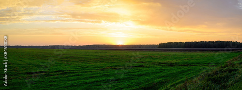 Panoramic photo of a green field in the orange setting sun in autumn against the backdrop of a dramatic sky