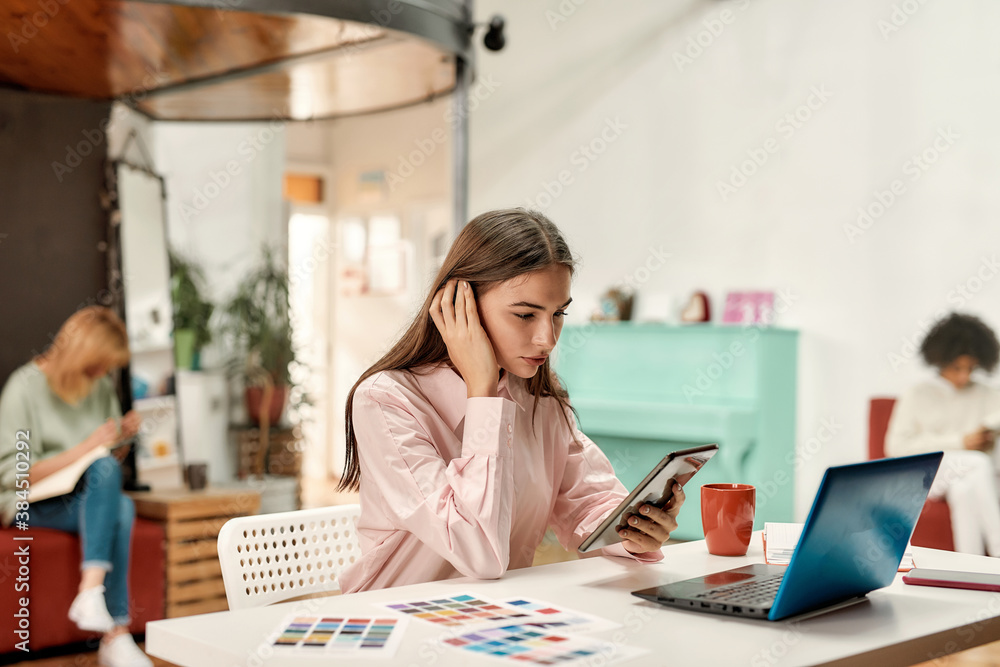 A cute young fashionably dressed woman with brown hair sitting at a table working with a tablet touching her hair A cute young fashionably dressed woman with brown hair sitting at a table working with a tablet touching her hair