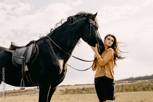 Young woman hugging a black friesian horse, outdoors, in a field, on windy weather.