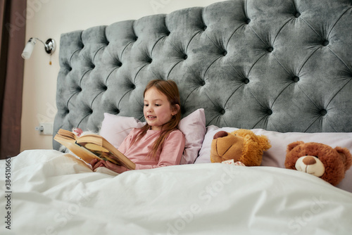 Fotografie A cute small girl half-lying on pillows in a huge bed covered with blanket readi