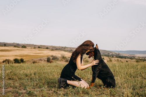 Young woman sitting on green grass, hugging a doberman breed dog, touching noses.