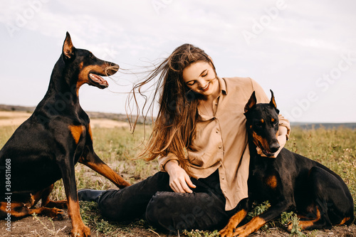 Print op canvas Young woman sitting on green grass with two doberman breed dogs.