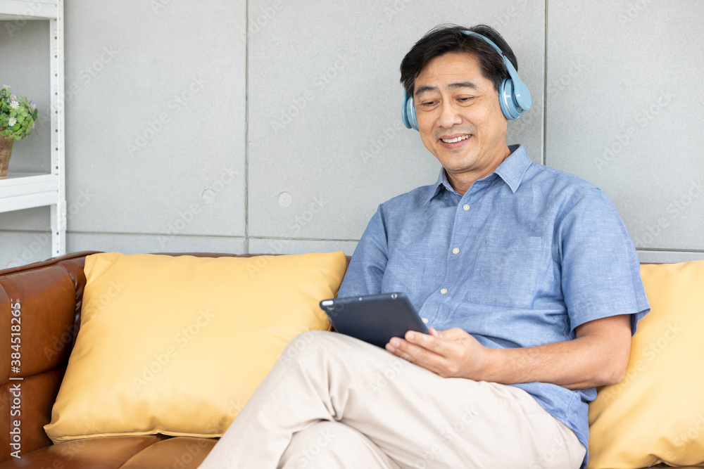 senior asian man wearing headphones and enjoy favorite music from tablet on sofa