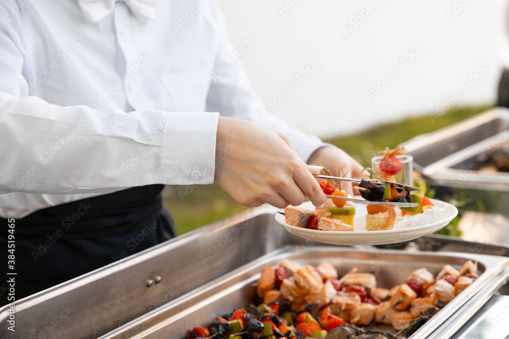 Outdoor catering banquet in summer. Waiter in white shirt puts a barbeque from a chafing dish on a plate. The waiter serves guests at the banquet