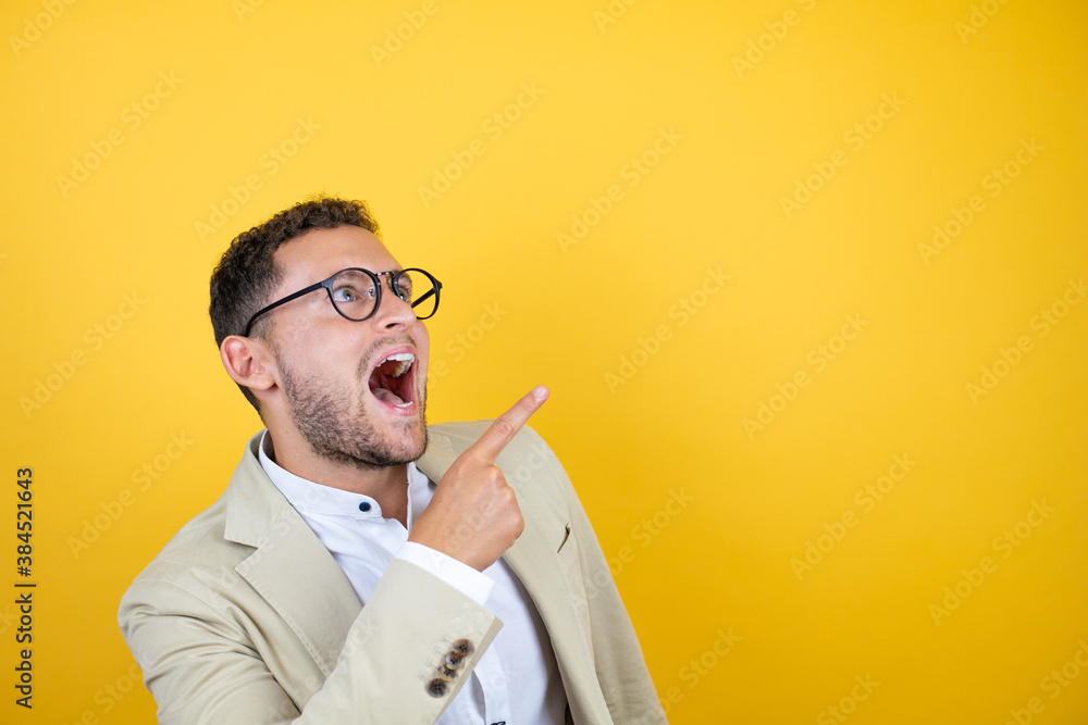 Young handsome businessman wearing suit over isolated yellow background surprised and pointing with hand and finger to the side Young handsome businessman wearing suit over isolated yellow background surprised and pointing with hand and finger to the side