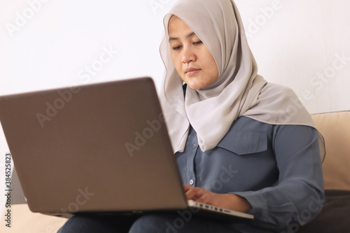 Asian muslim woman wearing hijab smiling while typing on her laptop, work from home concept. Young successful female entrepreneur working at home