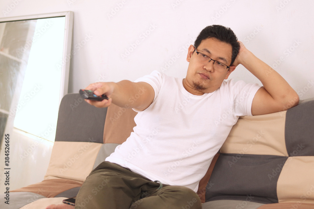 Fototapeta premium Bored tired young Asian man sitting on the sofa and watching TV. Trying to choose interesting TV channel
