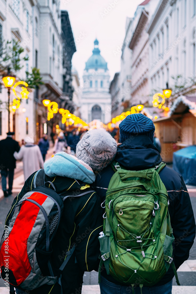A couple of tourists with backpacks walk the streets of Budapest, decorated for Christmas, against the background of St. Stephen's Basilica.