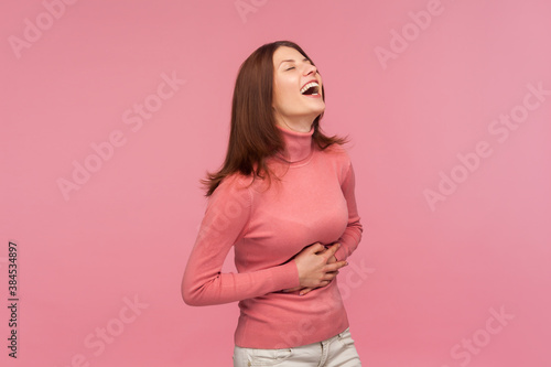 Funny positive brunette woman in pink sweater holding her belly and laughing out loud, excited careless female joking. Indoor studio shot isolated on pink background