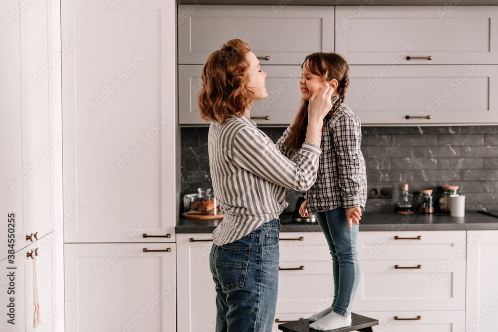 Little girl in jeans stands on chair and laughs. Mom touches daughters cheeks Little girl in jeans stands on chair and laughs. Mom touches daughters cheeks
