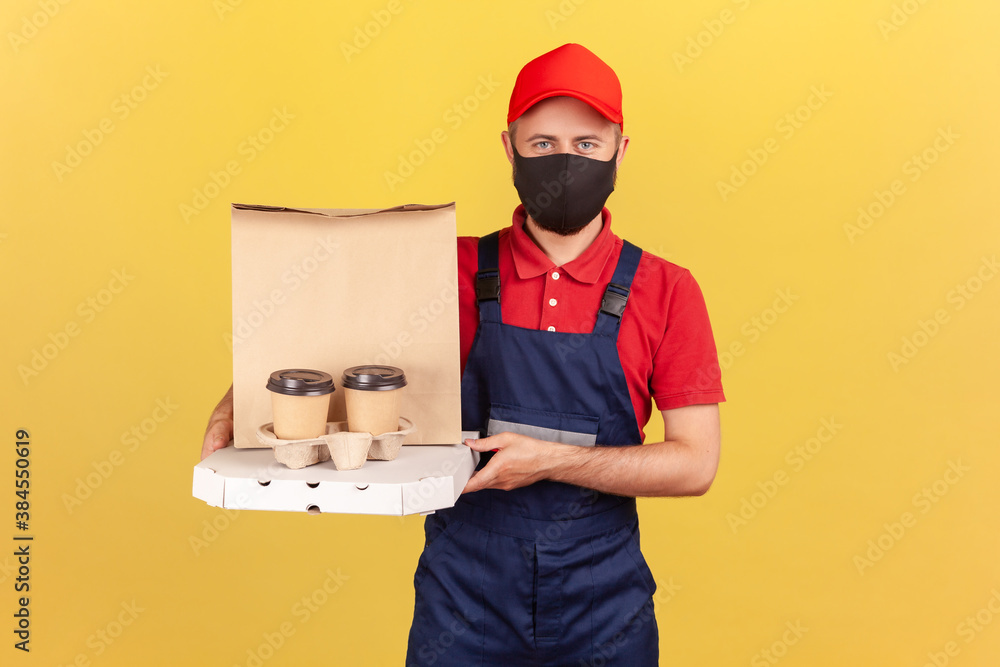 Delivery man in uniform and black medical protective mask holding coffee and boxes with pizza offering drinks and food delivery on quarantine. Indoor studio shot isolated on yellow background Delivery man in uniform and black medical protective mask holding coffee and boxes with pizza offering drinks and food delivery on quarantine. Indoor studio shot isolated on yellow background