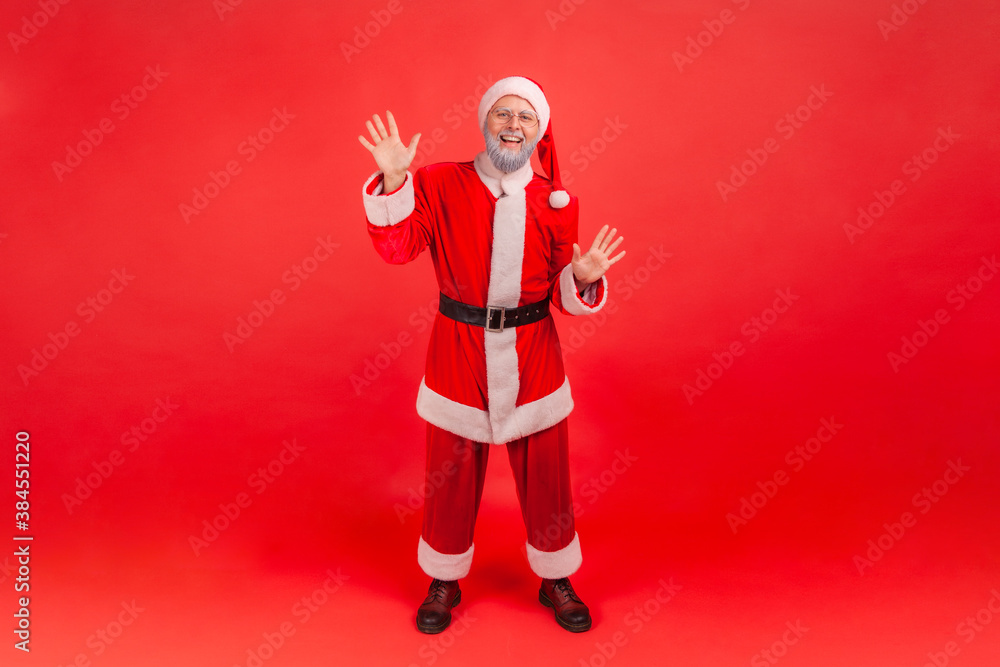 Full length portrait of friendly smiling gray bearded man in santa claus costume raising hands greeting and welcoming looking at camera. Indoor studio shot isolated on red background Full length portrait of friendly smiling gray bearded man in santa claus costume raising hands greeting and welcoming looking at camera. Indoor studio shot isolated on red background