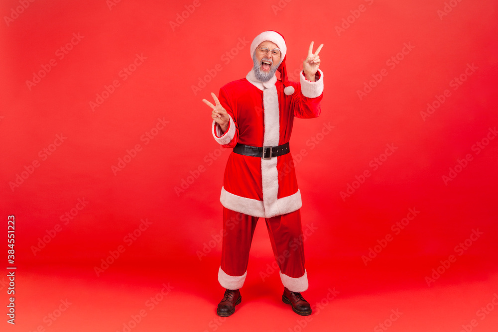 Full length portrait of positive funny gray bearded man in santa claus costume winking showing peace gesture with fingers, free and optimistic waiting for holidays. Indoor isolated on red background Full length portrait of positive funny gray bearded man in santa claus costume winking showing peace gesture with fingers, free and optimistic waiting for holidays. Indoor isolated on red background