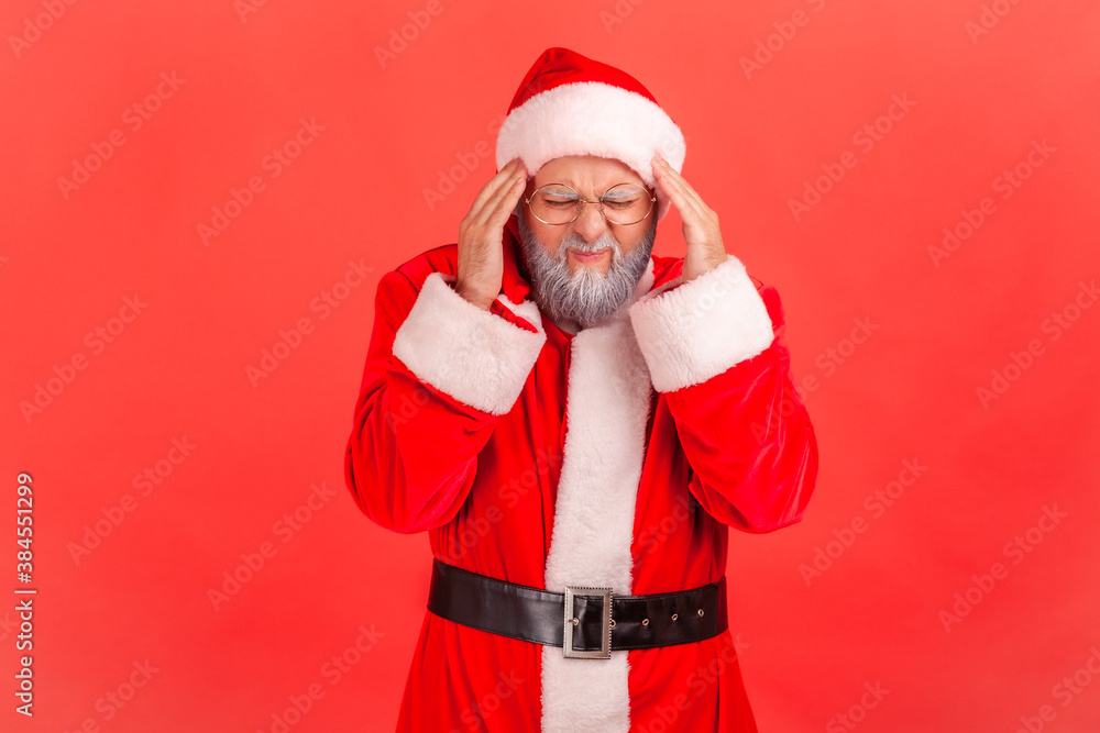 Portrait of sick elderly gray bearded santa claus massaging temples feeling unpleasant headache, risk of migraine, high blood pressure. Indoor studio shot isolated on red background