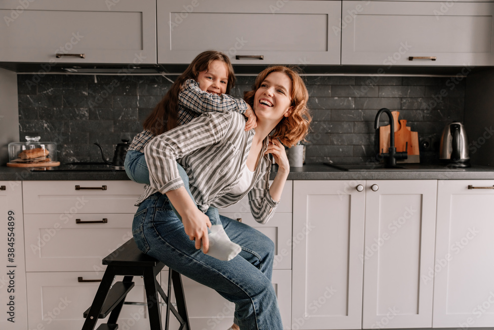 Beautiful woman in jeans sits on chair in kitchen while her daughter hugs her from behind Beautiful woman in jeans sits on chair in kitchen while her daughter hugs her from behind