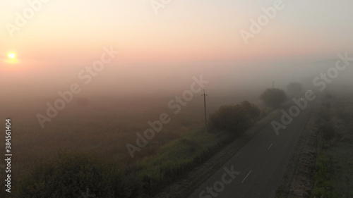 Foggy road near the field. Asphalt driveway covered by dense mist. Cars are riding in white haze.