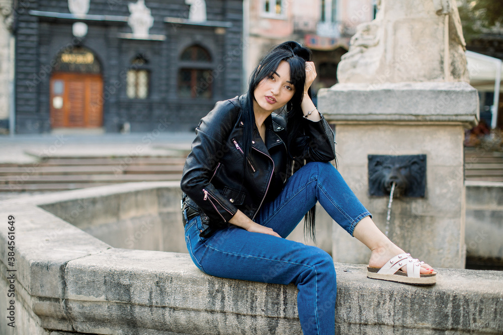 Beautiful young dreamy sexy Asian woman in trendy casual clothes, posing while sitting on stone fountain at old European city street and looking at camera. Travel, lifestyle, urban concept