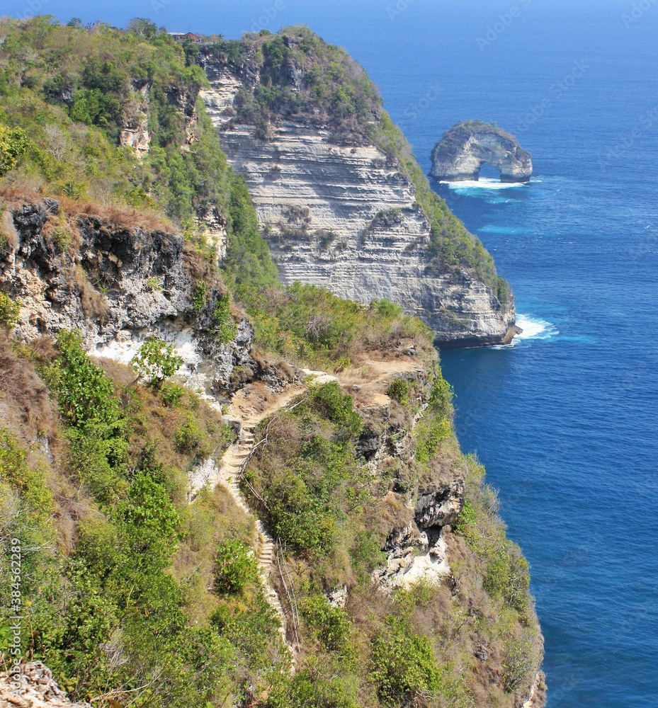 Steep cliff with rocky coastline, natural arch in background on Nusa Penida, Bali, Indonesia