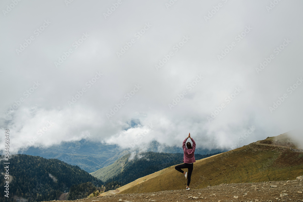 Asana high up in cloudy mountains. Young woman doing yoga stance. Long shot