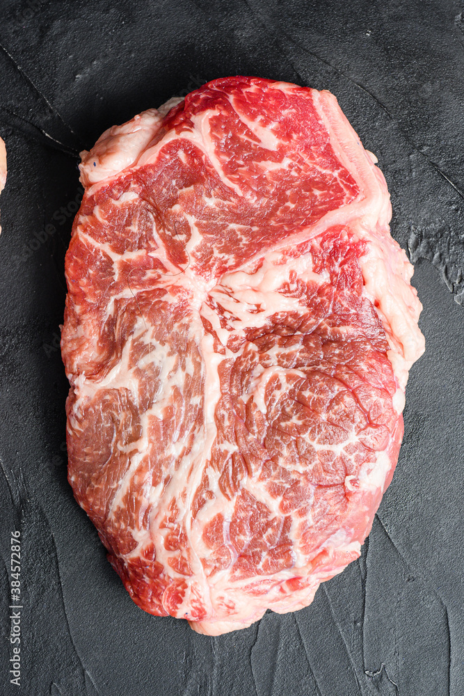 Raw top blade beef steak cut, on black textured background, top view.