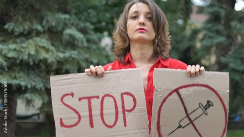 Young protesting woman in red shirt holds protest sign broadsheet placard with slogan 'Stop vaccine' for public demonstration on trees background.