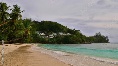 Tourists enjoying their holidays on tropical beach in Baie Lazare, Mahe island, Seychelles with turquoise colored water, coconut trees, docking boats and holiday homes in the background.